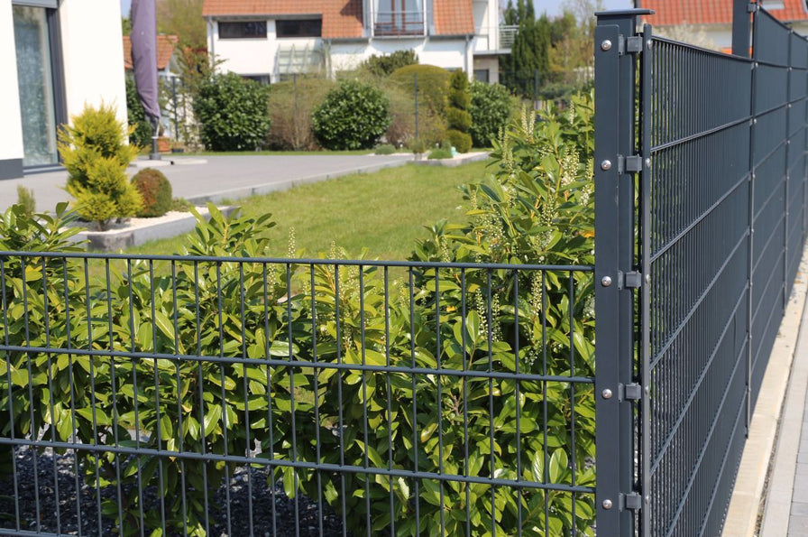 Anwendungsbild eines Doppelstabmattenzauns in der Ausführung 6/5/6 mit einem Eck-Zaunpfosten und weiteren Zaunpfosten, jeweils mit Abdeckleiste. Im Hintergrund sind Rasenfläche, Haus und Terrasse zu sehen.