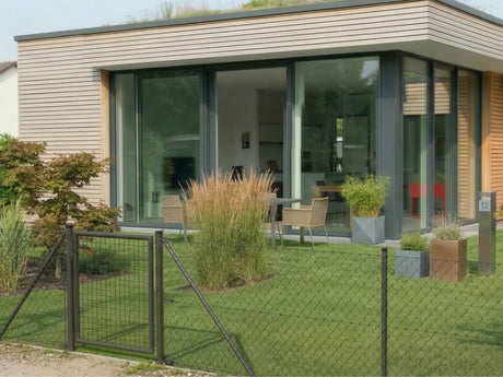 Ein Maschendrahtzaun in der Farbe anthrazit. Der Zaun steht vor einem modernen Haus mit Garten und dient als Abgrenzung des Grundstuecks.