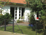 Ein Maschendrahtzaun mit Tor in der Farbe anthrazit, der als Abgrenzung von einem Grundstück dient. Im Hintergrund ist ein kleiner Garten mit Haus abgebildet